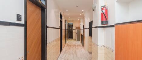 A hotel corridor with a fire extinguisher mounted on the wall, a door on the left, and a hallway leading to a room.