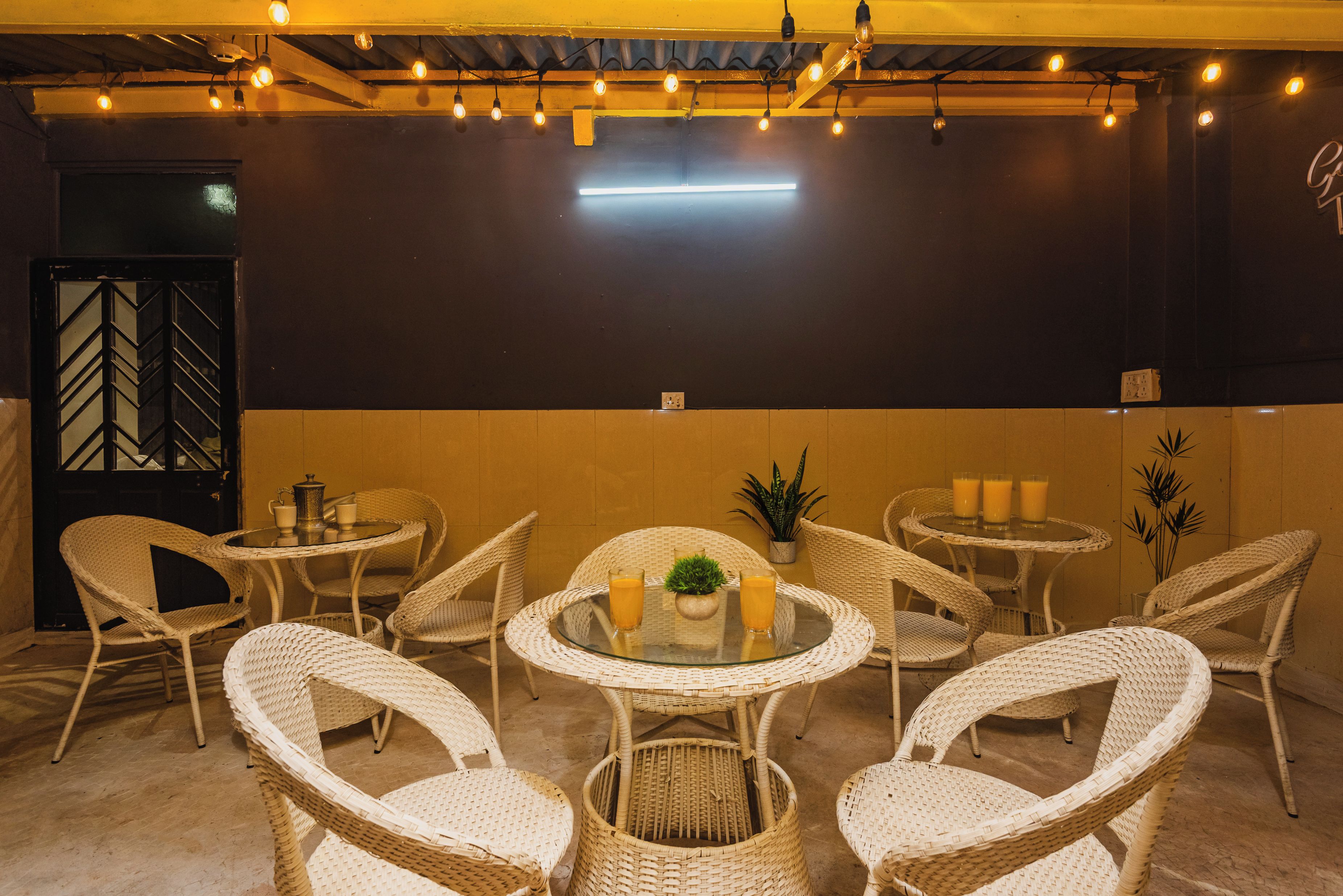 A cozy outdoor seating area with wicker chairs and tables, overhead string lights, and a wall-mounted light fixture.