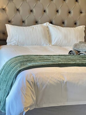 A bed with white bedding and a green blanket, a tufted headboard, and a rolled-up grey blanket on the side. - Luxury Apartment Overlooking the River (Midrand)