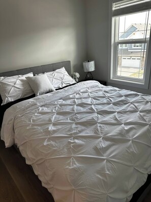 A neatly made bed with a white duvet and pillows, a bedside lamp, and a window with a view of a house. - Luxury Home in Aspen (Calgary)