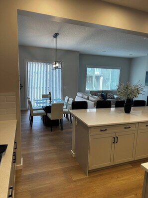 A modern kitchen with white cabinetry, a dining area with a glass table, and a living room with a sofa and a potted plant. - Luxury Home in Aspen (Calgary)