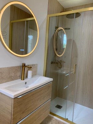 Un baño con un espejo redondo, un lavabo blanco, un grifo dorado y una ducha con cerramiento de vidrio.