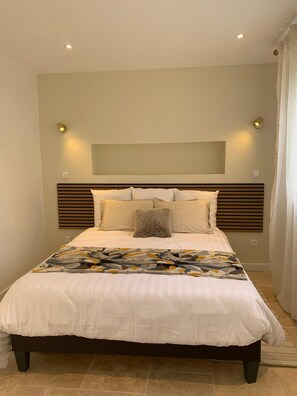Un dormitorio con cama, cabecera, lámparas de noche y una ventana con cortinas.