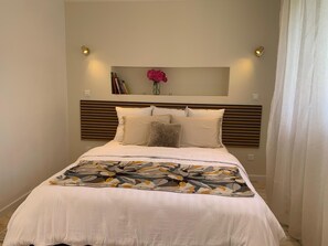 Een keurig opgemaakt bed met een bloemenprint plaid, een houten hoofdbord en een wandplank met boeken en een bloevas.