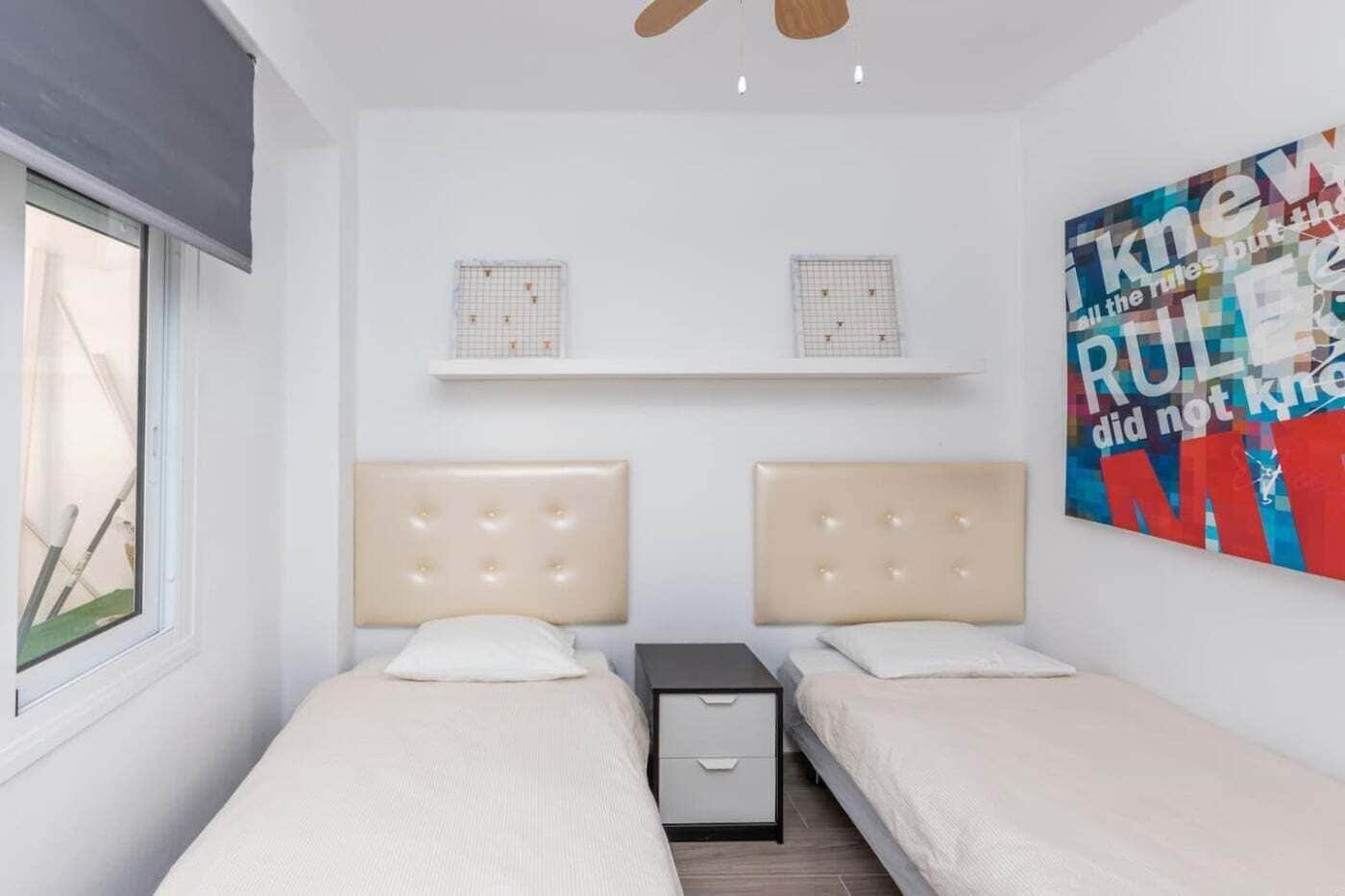 A double room with two beds, a small bedside table, a nightstand, a ceiling fan, and a wall-mounted shelf with two decorative items.