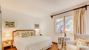 A bedroom with a bed, a chair, a nightstand with a lamp, a table with a lamp, and a window with curtains. - Beautiful House with Wi-Fi, Air Conditioning, and Pool in Exclusive Community (Cala de Sant Vicenç)