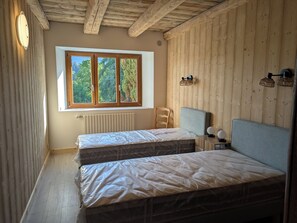 A room with two beds, wooden walls, a window with a view of trees, a radiator, and a ceiling with exposed beams. - Large T4 garden apartment in a renovated farmhouse in a peaceful village. (Mieussy)