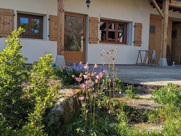 A building with wooden shutters, a stone foundation, and a garden with various plants and flowers. - Large T4 garden apartment in a renovated farmhouse in a peaceful village. (Mieussy)