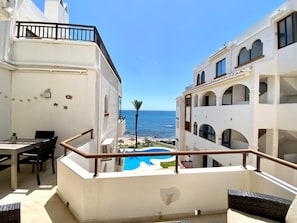A balcony with a view of the sea, a pool, and a dining area with chairs and a table. - Beachfront 2BR scenic sea views - Pool (Mijas)