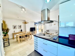 A modern kitchen with a central island, a dining area with wooden furniture, and a living space with a sofa and a TV. - Beachfront 2BR scenic sea views - Pool (Mijas)