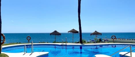 Una piscina a curvatura sinuosa, circondata da palme e ombrelloni da spiaggia, con l'oceano sullo sfondo.