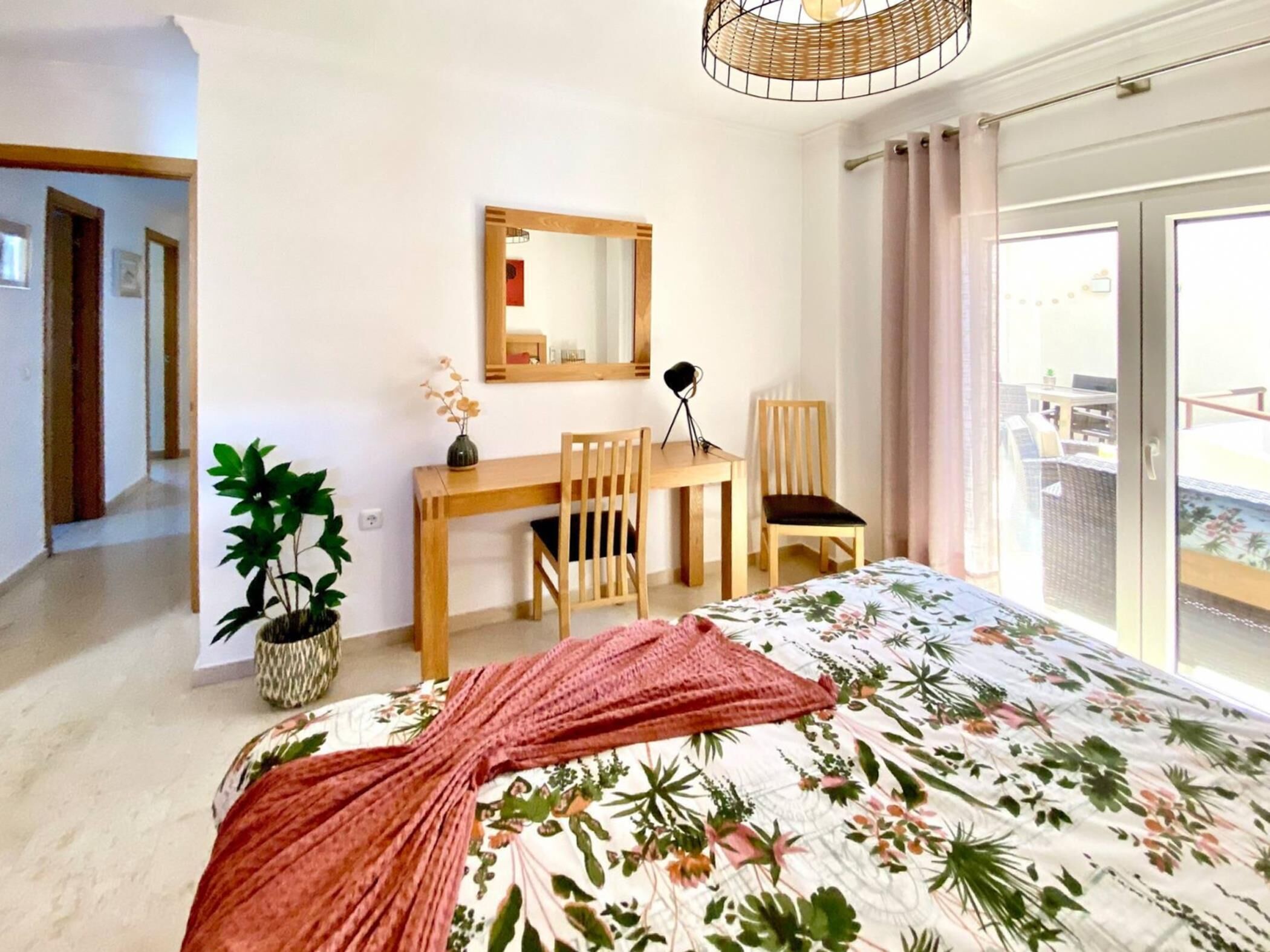 Un dormitorio con cama, escritorio, dos sillas, espejo, planta en maceta y vista al balcón.