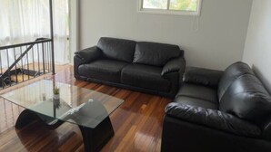 A living room with a glass coffee table, two black leather sofas, wooden flooring, a ceiling fan, and a window with natural light. - Riverview Retreat 3 BDR house 2 toilets (Riverview)