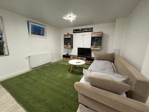 A living room with a green carpet, a beige sofa, a TV, and built-in shelves. - Business Apartment Kaiserslautern · Sleeps 2 · Full Kitchen · Living Room (Kaiserslautern)