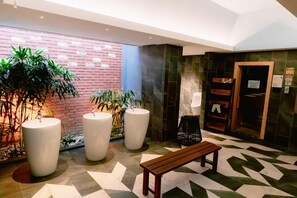 A lobby with a brick wall, large potted plants, a wooden bench, and a reception desk.