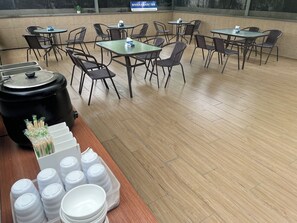 A restaurant with multiple tables and chairs, a large black container, and a table with stacked white bowls and lids.