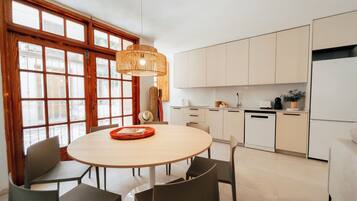 Een moderne keuken met witte kasten, een ronde eettafel met stoelen en een groot raam met houten kozijnen.