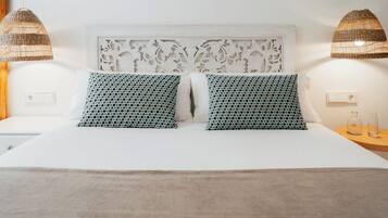 Un lit avec une tête de lit blanche et deux coussins à motifs, deux lampes fixées au mur et une table de chevet avec un récipient en verre et une bouteille.