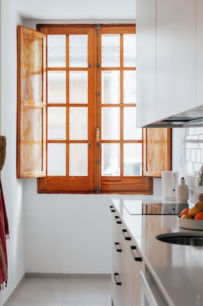 A kitchen with a large wooden-framed window, white cabinets, and a countertop with a coffee maker and fruit bowl.