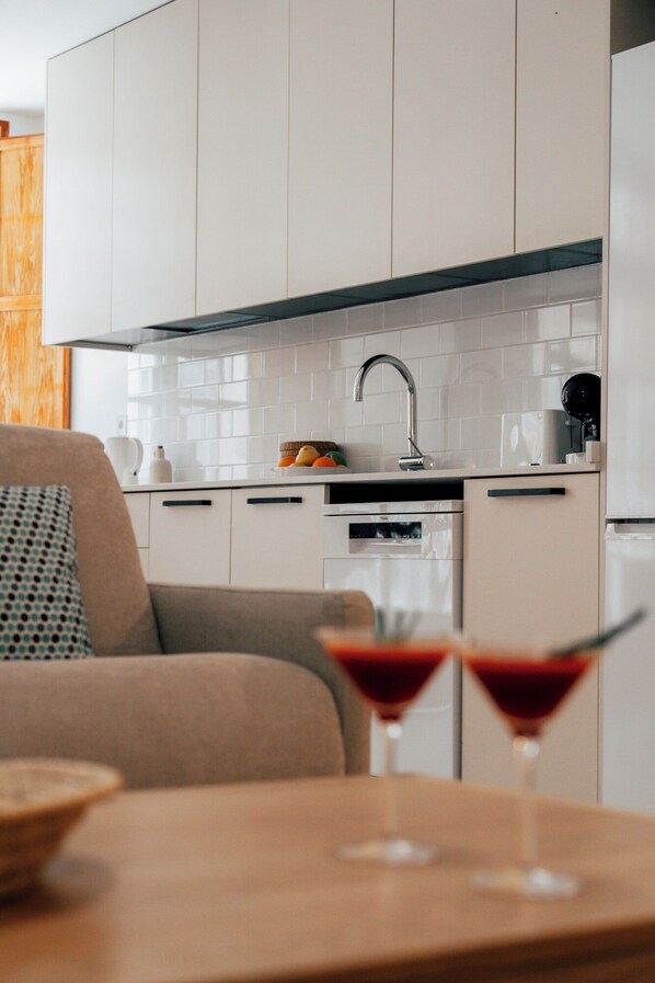 A modern kitchen with white cabinets, a black countertop, and a built-in oven. There is a wooden table with two martini glasses in the foreground.