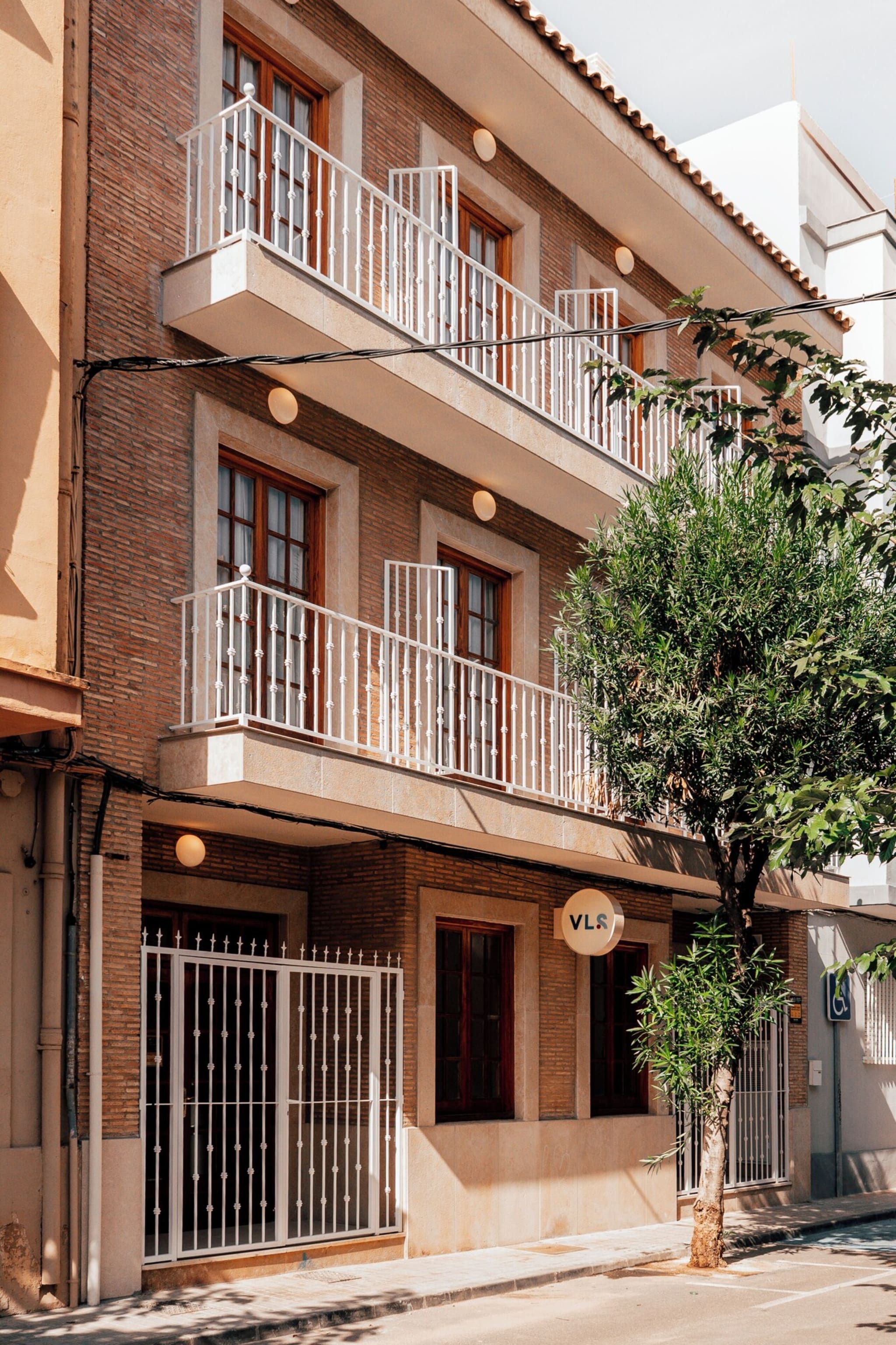 Un immeuble à plusieurs étages, doté d’une façade en briques, de balcons avec des garde-corps blancs et d’un arbre devant.