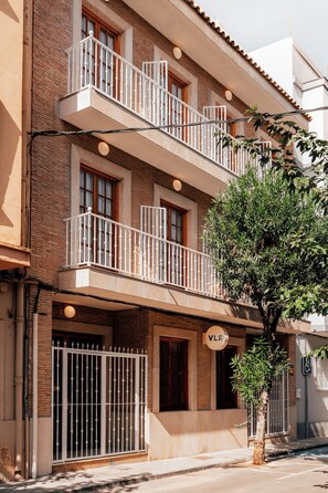 A multi-story building with a brick facade, balconies with white railings, and a tree in front.