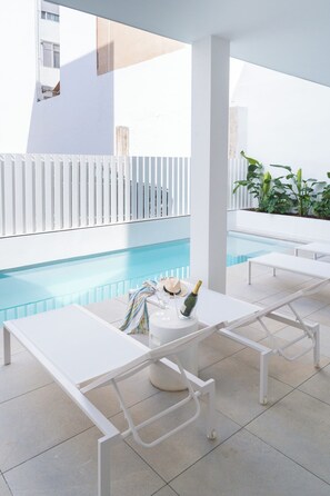 A poolside area with white lounge chairs, a table with a bottle of champagne, and a tiled floor.