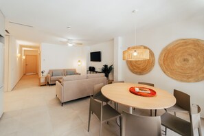 Una sala moderna con un sofá, una mesa de comedor y sillas, un televisor montado en la pared y un pasillo que lleva a otra habitación.