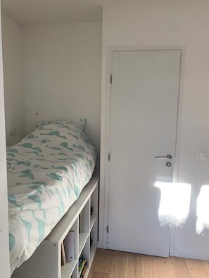 Een kleine kamer met een enkel bed, een witte deur, een planken kast onder het bed en een wandlamp.