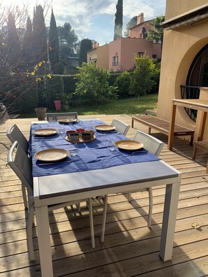 A wooden deck with a table set for a meal, surrounded by chairs, and a view of a garden and buildings in the background. - Vrbo Property (Cagnes-sur-Mer)