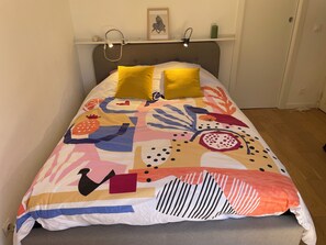 A bed with a colorful abstract bedspread, two pillows, a headboard, and a shelf with a framed picture and a green candle. - Vrbo Property (Cagnes-sur-Mer)