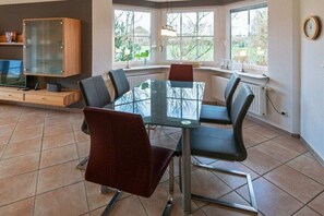 A dining area with a glass table, four chairs, a cabinet, and large windows. - Holiday home with 3 bedrooms (Norddeich)