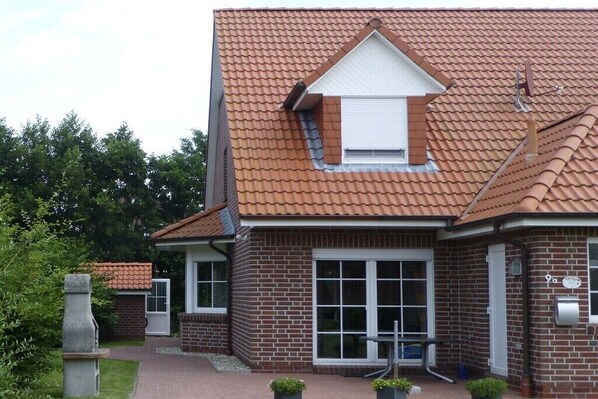 A two-story brick house with a red-tiled roof, a small attached garage, and a front porch with potted plants. - Holiday home with 3 bedrooms (Norddeich)