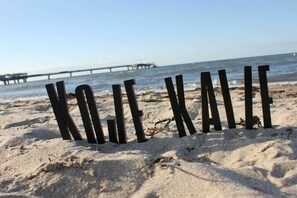 Letras de madera que deletrean 'KOJI XAVI' sobre una playa arenosa, con un muelle al fondo.