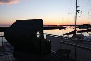 Eine Terrasse mit einem Korbstuhl, einem kleinen Tisch und Blick auf den Yachthafen mit Booten und einem Sonnenuntergang.