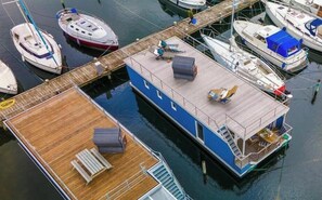 Une péniche flottante est amarrée dans une marina. Elle est dotée d’une terrasse en bois, d’un espace salon extérieur et d’un petit escalier menant à la terrasse supérieure.