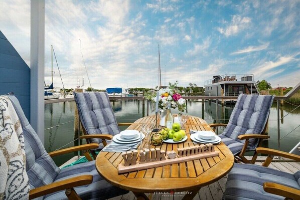 Une terrasse en bois avec deux chaises à rayures, une table ronde mise avec des assiettes, des verres et un centre de table composé de fruits.