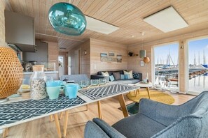 Un interno moderno con soffitto in legno, un'area pranzo con tavolo decorato, un divano e vista sulle barche attraverso ampie finestre.