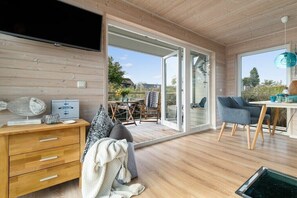 Un intérieur moderne avec un plancher en bois, un téléviseur fixé au mur, un coin repas avec une table et des chaises, et une grande porte-fenêtre coulissante donnant sur un espace extérieur.