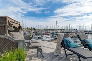 Une terrasse dotée de chaises longues et d’un fauteuil en osier, offrant une vue sur une marina où sont amarrés de nombreux bateaux.
