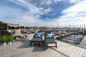 Une terrasse en bois avec des chaises longues et un canapé, donnant sur une marina où sont amarrés de nombreux bateaux.