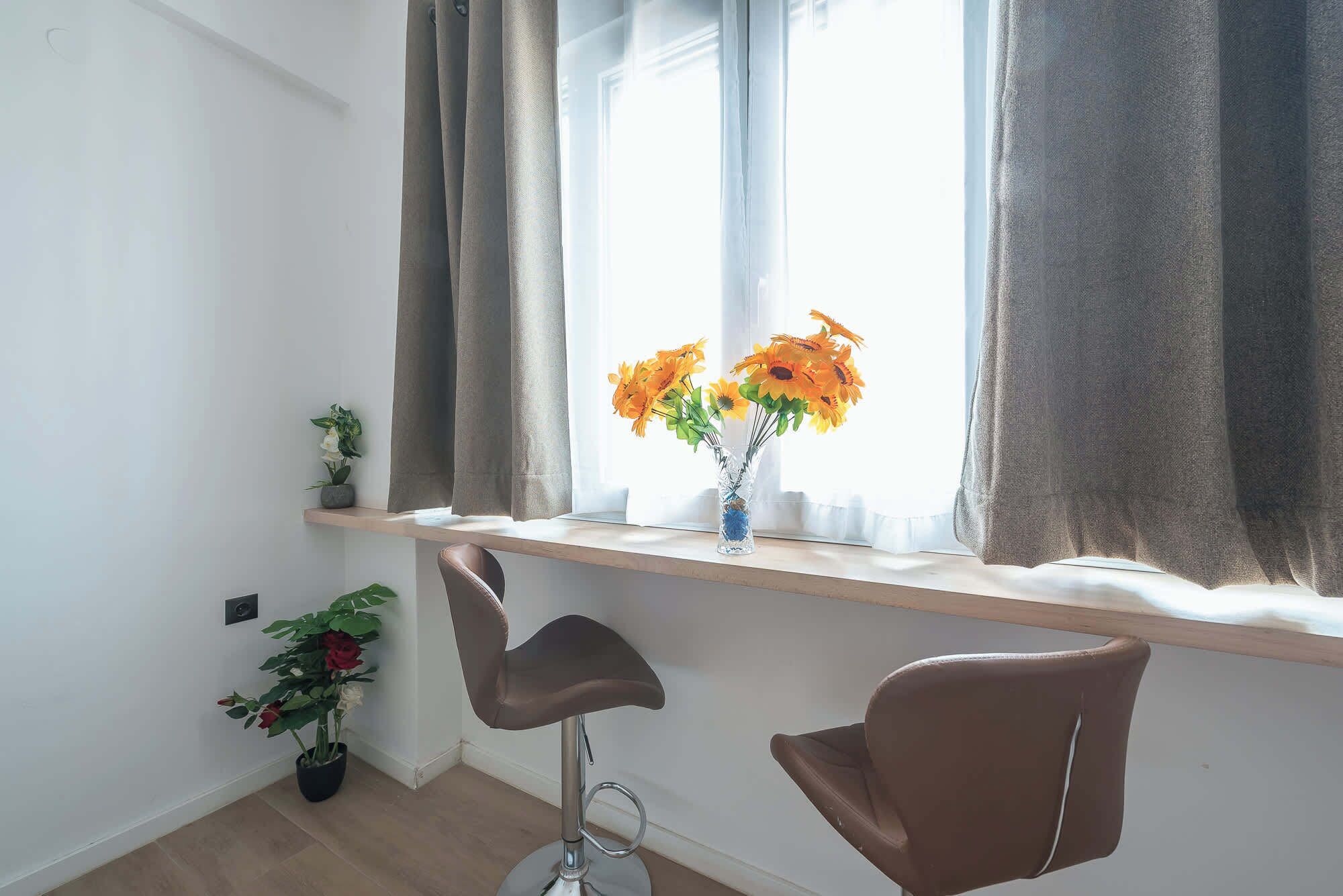 Una habitación con ventana, un jarrón con flores, una planta en maceta y dos sillas.