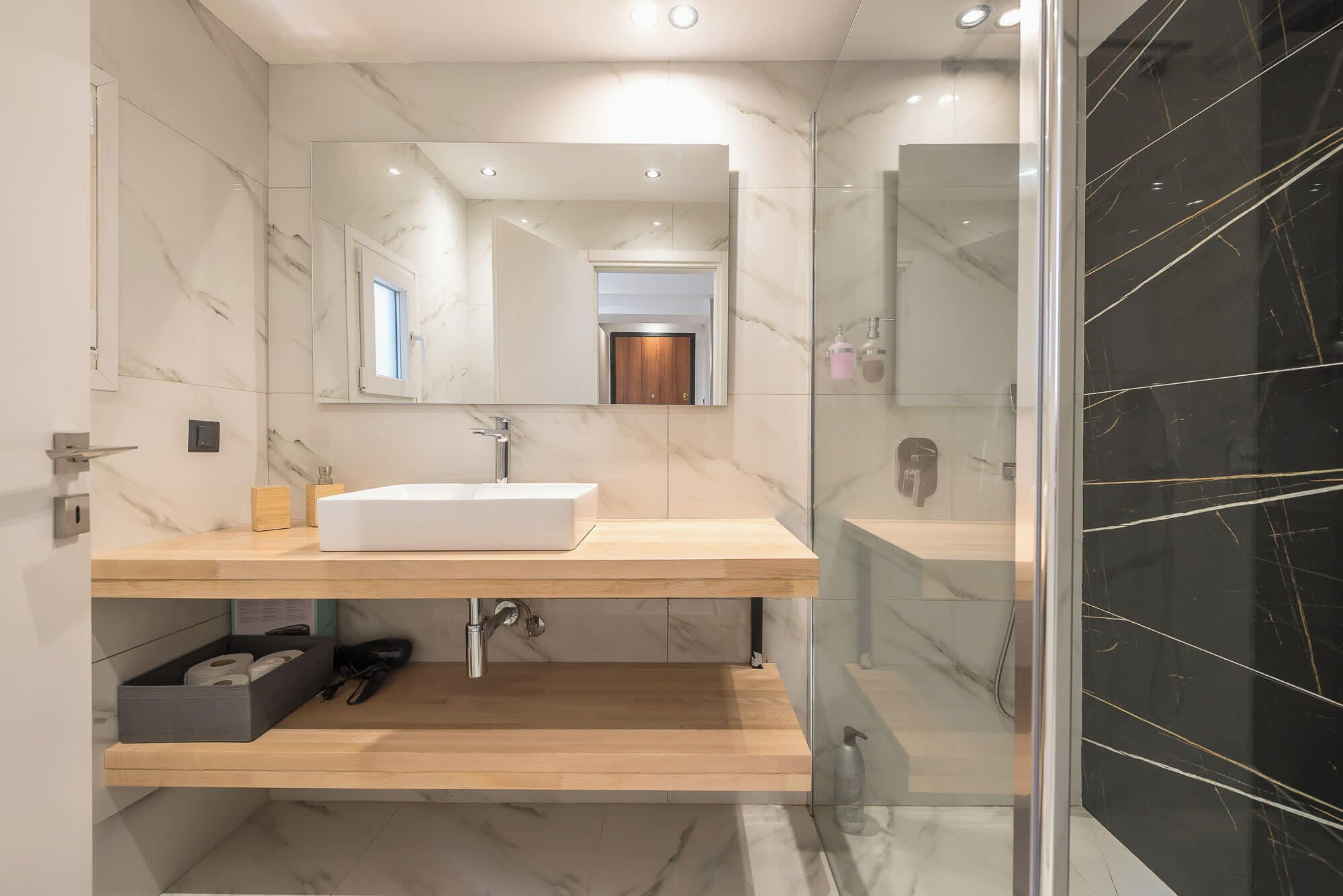 A modern bathroom with a white sink, wooden vanity, marble walls, and a glass shower enclosure.