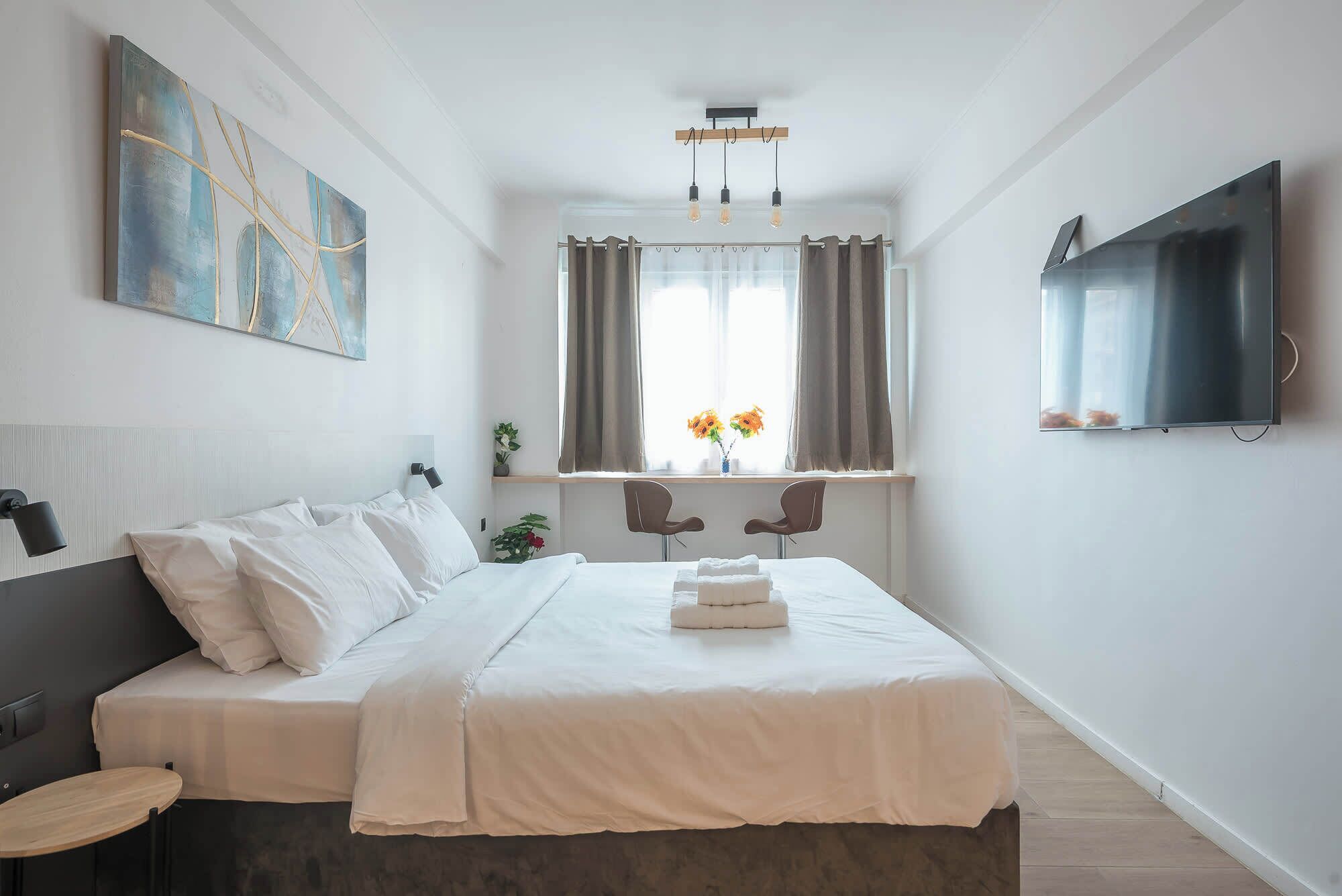 A modern hotel room with a bed, a TV, a painting, a window with curtains, and a plant.