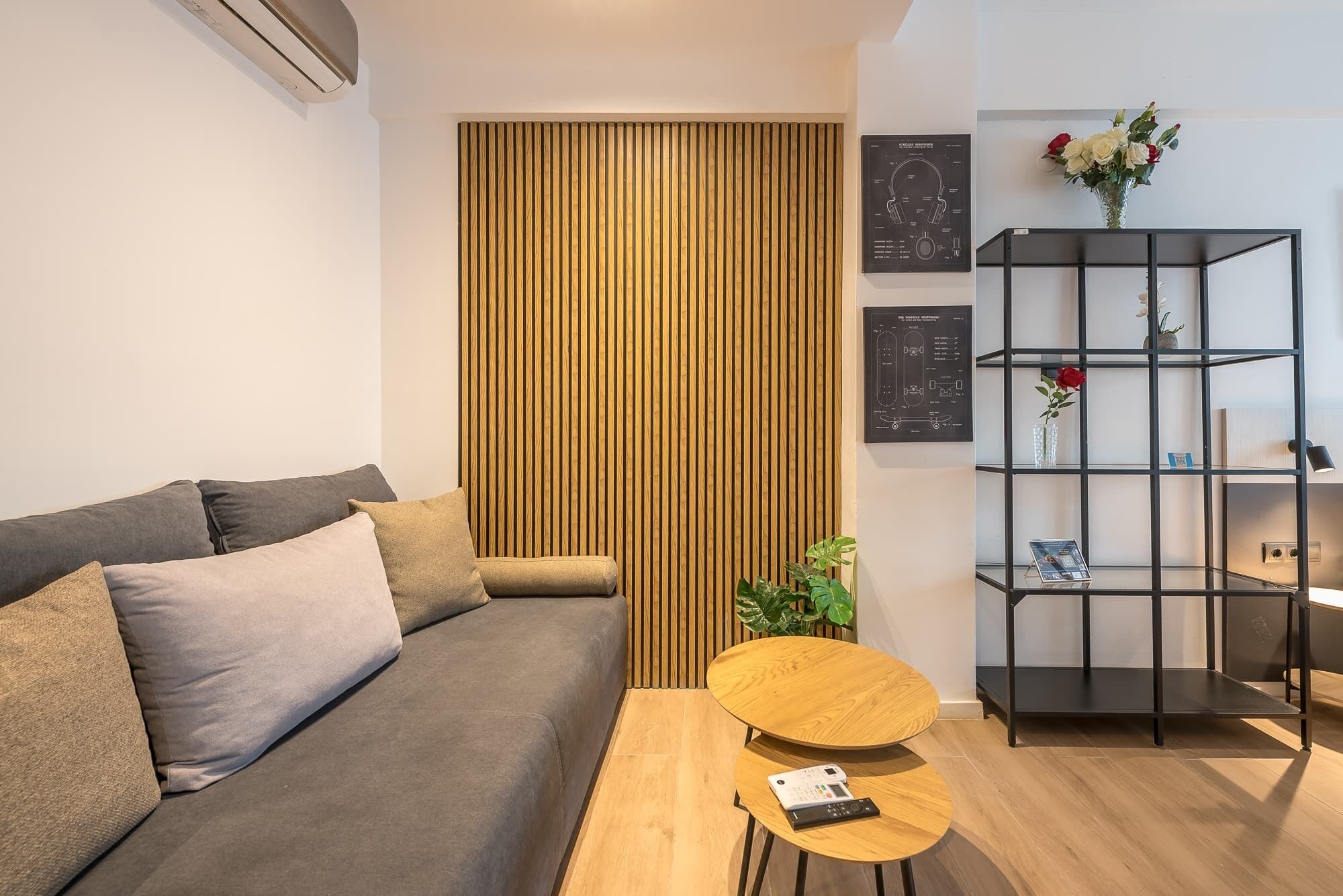 Un salon moderne avec un canapé gris, une cloison en lattes de bois, une étagère noire agrémentée d’objets décoratifs et une petite table ronde en bois.