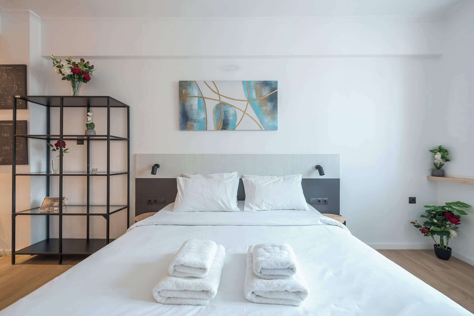 Ein Hotelzimmer mit einem Bett, zwei Nachttischen, einem an der Wand befestigten abstrakten Gemälde, einer schwarzen Regaleinheit mit Blumen und einer Topfpflanze.