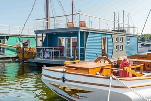 Een klein, blauw woonschip afgemeerd in een jachthaven, met een houten dek en een stuurwiel.