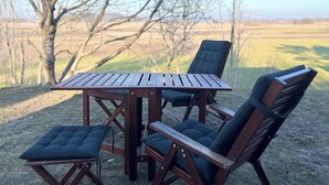 Una mesa de madera y cuatro sillas con asientos acolchados, ubicadas al aire libre sobre un suelo de grava, con un árbol y un campo abierto al fondo.