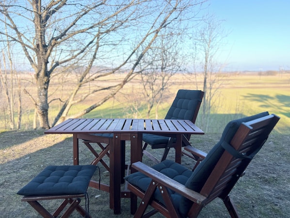 Una mesa de madera y cuatro sillas con asientos acolchados, ubicadas al aire libre sobre un suelo de grava, con un árbol y un campo abierto al fondo.