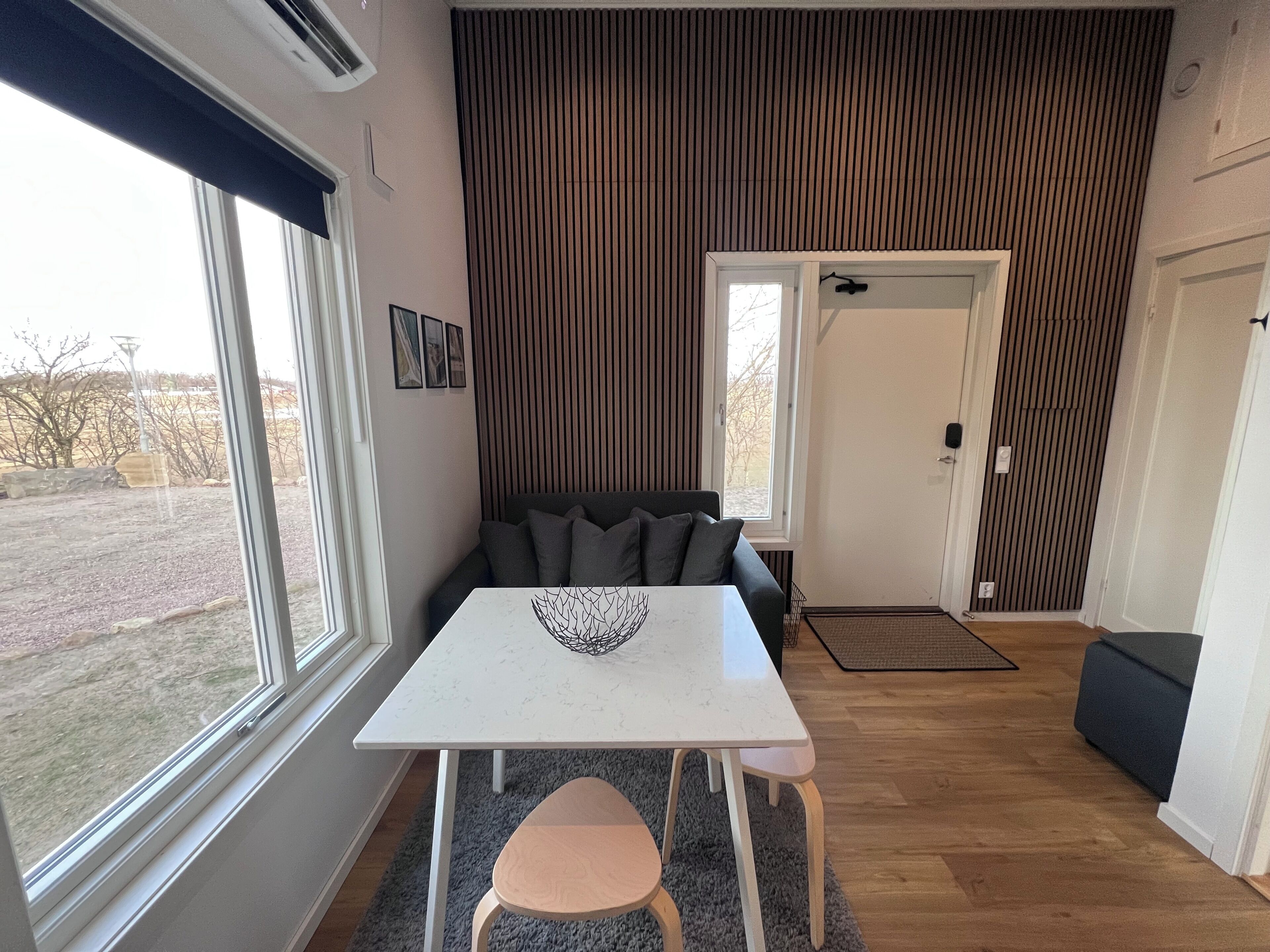 Ett modernt vardagsrum med ett vitt bord, en grå soffa och utsikt över naturen genom stora fönster.